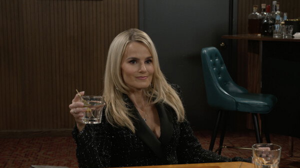 A medium shot of a blonde woman with long, straight hair, looking directly at the camera with a slight, confident smile. She is holding a martini glass, garnished with a toothpick, in her raised right hand. She is wearing a black, sparkly jacket with satin lapels. The background features a dark wood-paneled wall, a teal tufted bar stool, and a small table holding various liquor bottles, suggesting a bar or upscale lounge setting. The lighting is relatively dim and dramatic, contributing to a chic and poised mood, likely from a television show or film.