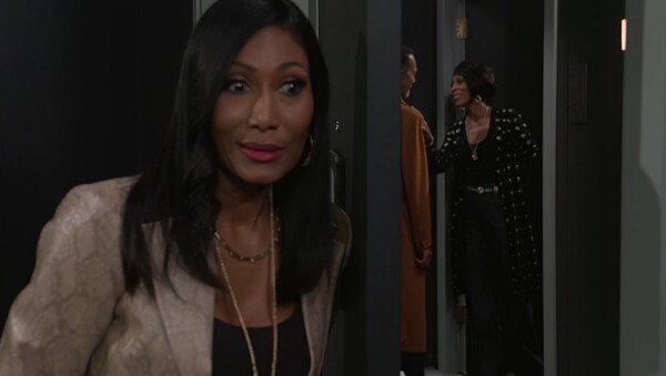 A cinematic image featuring three people in a doorway setting. In the foreground, a Black woman with long dark hair, wearing a snakeskin-patterned blazer and several gold necklaces, is seen looking to the side with a surprised or intense expression. Her face is slightly blurred as if she is moving quickly or peaking around a corner. In the background, framed within a dark doorway, two other people are interacting: a person in an orange outfit, seen mostly from the back, and a woman in a black outfit with white polka dots or studs, who is smiling toward the person in orange. The lighting is low-key and dramatic, suggesting a moment of confrontation or secret observation from a television show or movie.