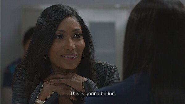 A medium shot of an attractive Black woman with long, dark hair, wearing a black leather jacket, looking off-camera to her right with a subtle, knowing smile. She rests her chin on her clasped hands. The scene is indoors, brightly lit, suggesting a television show or movie still, which is confirmed by the subtitle at the bottom that reads: 'This is gonna be fun.' The overall mood is confident and slightly conspiratorial.