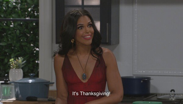 A medium close-up of a woman with medium-brown skin and dark, wavy hair, wearing a plunging, deep red dress. She has a bright, excited smile and is looking up and slightly to her right. She wears a large gold pendant necklace with a blue stone and gold hoop earrings. The image is a still from a television show, and text overlaid at the bottom says, 'It's Thanksgiving!' She is standing in what appears to be a modern kitchen, with a black stove and light gray cabinets visible in the background, along with a teal Dutch oven on the counter to her left. The overall mood is joyful and festive, captured with bright, high-quality lighting typical of a TV production.