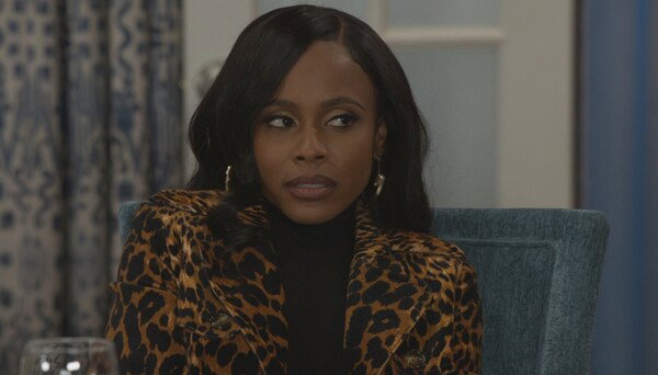 A medium close-up of a woman with dark brown skin and sleek, dark hair parted down the middle, sitting in a dining room setting. She is wearing a black turtleneck underneath a statement coat in a vibrant brown and black leopard print. She has a serious, slightly tense expression, looking intently off-camera to her left. She wears gold, angular hoop earrings. The background shows a light blue upholstered chair and partially visible dark blue and cream patterned curtains. The image is a still from a television show, characterized by clean, direct lighting and a focused, dramatic mood.