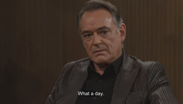 A medium close-up of an older man with light brown hair swept back, wearing a dark gray or black textured sport coat over a black button-up shirt. He has a serious, weary expression, looking directly at the camera. The background is a plain, dark brown wall with subtle vertical ribbing. The image is a still from a video or television show, with the text 'What a day.' overlaid at the bottom. The overall mood is somber and reflective, captured with low-key, dramatic lighting.
