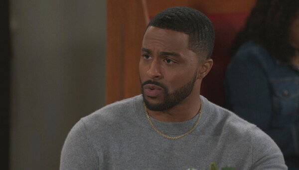 A medium close-up of a man with dark brown skin, a neatly trimmed beard, and short, dark hair with waves. He is wearing a plain, light gray crewneck sweater and a simple gold chain necklace. He has a serious, somewhat concerned expression, looking intently off-camera to his left. A portion of a woman's body in a dark blue denim jacket is visible and out of focus in the right background, with a dark wooden wall behind her. The image is a still from a television show, utilizing soft, even lighting and conveying a concerned or attentive mood.