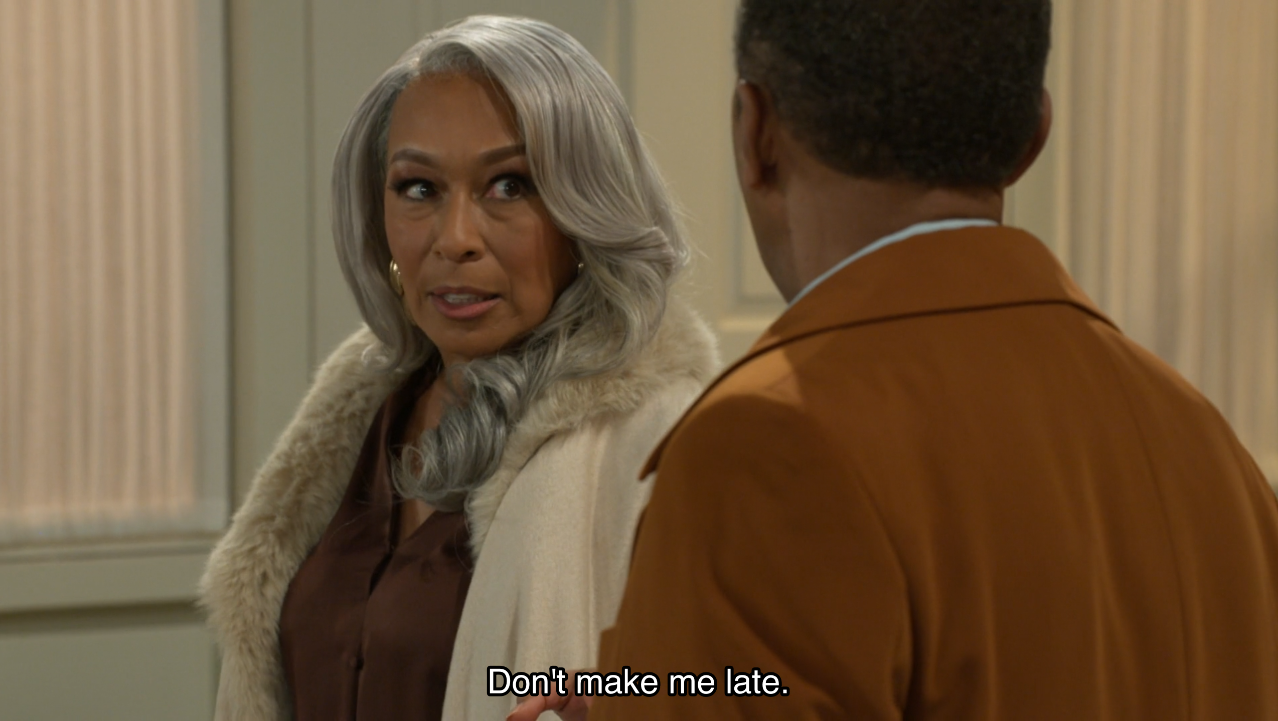 This cinematic film still features an elegant older woman with long, wavy silver hair and a cream-colored coat with a plush faux-fur collar, looking toward a man whose back is to the camera. She wears a brown blouse and gold hoop earrings, her expression one of firm expectation or slight impatience as she speaks. At the bottom of the frame, white subtitles read, "Don't make me late." The lighting is soft and warm, characteristic of a high-end television drama, set against a neutral, sophisticated interior that enhances the scene's mood of domestic urgency.