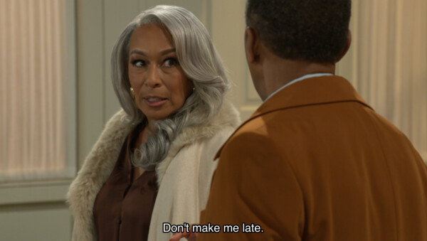 This cinematic film still features an elegant older woman with long, wavy silver hair and a cream-colored coat with a plush faux-fur collar, looking toward a man whose back is to the camera. She wears a brown blouse and gold hoop earrings, her expression one of firm expectation or slight impatience as she speaks. At the bottom of the frame, white subtitles read, 'Don't make me late.' The lighting is soft and warm, characteristic of a high-end television drama, set against a neutral, sophisticated interior that enhances the scene's mood of domestic urgency.