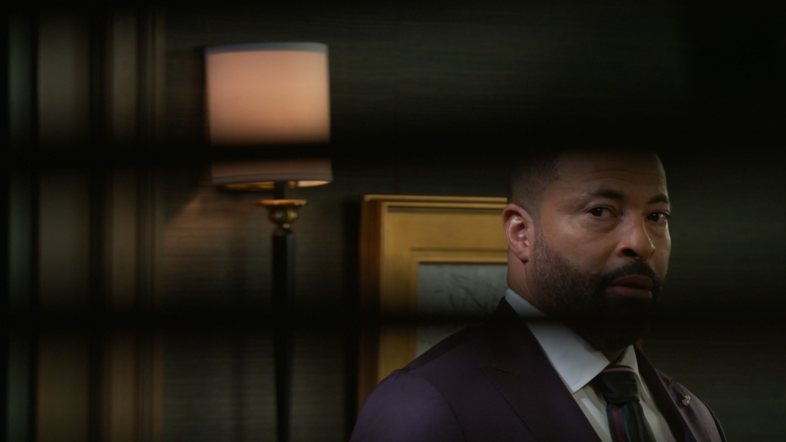 A medium close-up shot of a Black man with a groomed beard and a serious, concerned expression, dressed in a dark suit, white shirt, and striped tie. He is positioned in an elegantly dim room, with a soft-lit wall lamp and a gold-framed picture visible in the background. The entire scene is viewed through the dark horizontal slats of blinds, which create a cinematic, voyeuristic perspective. The mood is tense and dramatic, suggesting a moment of suspense or observation within a high-stakes professional or legal environment.
