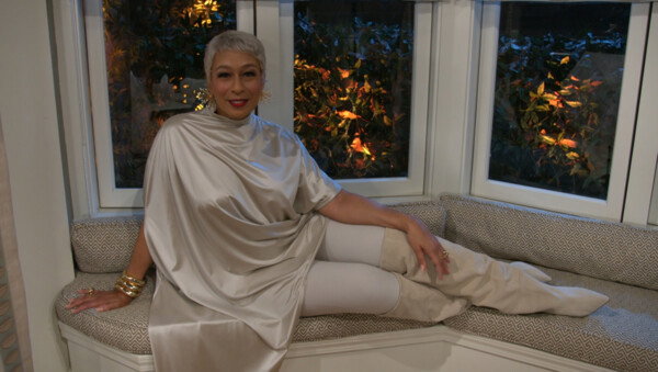 A woman with short, silver hair and a sophisticated smile reclines on a patterned window seat, dressed in a shimmering, champagne-colored draped tunic, light-colored leggings, and tall suede boots. She is adorned with bold gold statement jewelry, including large earrings and stacked bangles, which complement the warm lighting of the indoor setting. Behind her, a large bay window reveals a glimpse of lush green foliage illuminated by soft outdoor lights at twilight. The overall style is elegant and luxurious, conveying a sense of high-fashion comfort and mature grace.