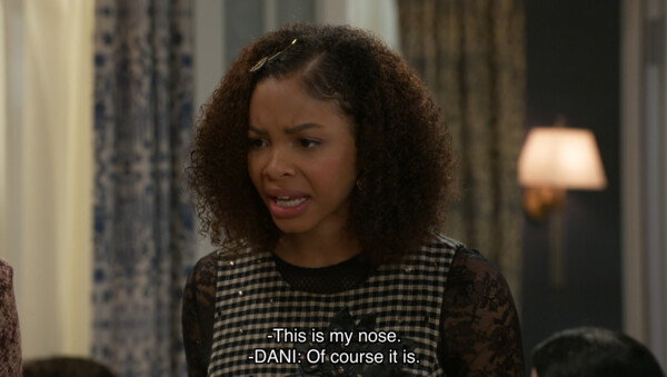 A medium shot captures a young woman with curly brown hair and a skeptical, furrowed expression during a scene from a television comedy. She is wearing a black lace-sleeved top under a black-and-white checkered vest and has a small decorative clip in her hair. The background consists of a softly lit interior with patterned curtains and a warm lamp, characteristic of a multi-camera sitcom set. Overlaid at the bottom of the frame are white subtitles that read, '-This is my nose. -DANI: Of course it is,' suggesting a moment of dry humor or an absurd argument. The overall mood is lighthearted yet tense, leaning into the expressive, character-driven style of a modern sitcom.