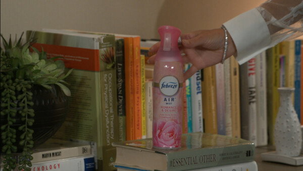 A person's hand with a silver bracelet and white lace sleeve reaches for a pink bottle of Febreze Air Mist in 'Romance & Desire' scent, which sits atop a stack of hardcover books on a shelf. The background features a diverse collection of books, including titles like Occupational Therapy for Physical Dysfunction and Life-Span Development, alongside a small green succulent in a dark textured pot and a white decorative vase. The overall mood is soft, domestic, and studious, utilizing warm, natural lighting and a shallow depth of field to create a cozy, lived-in atmosphere that suggests a moment of personal care or home refreshing.