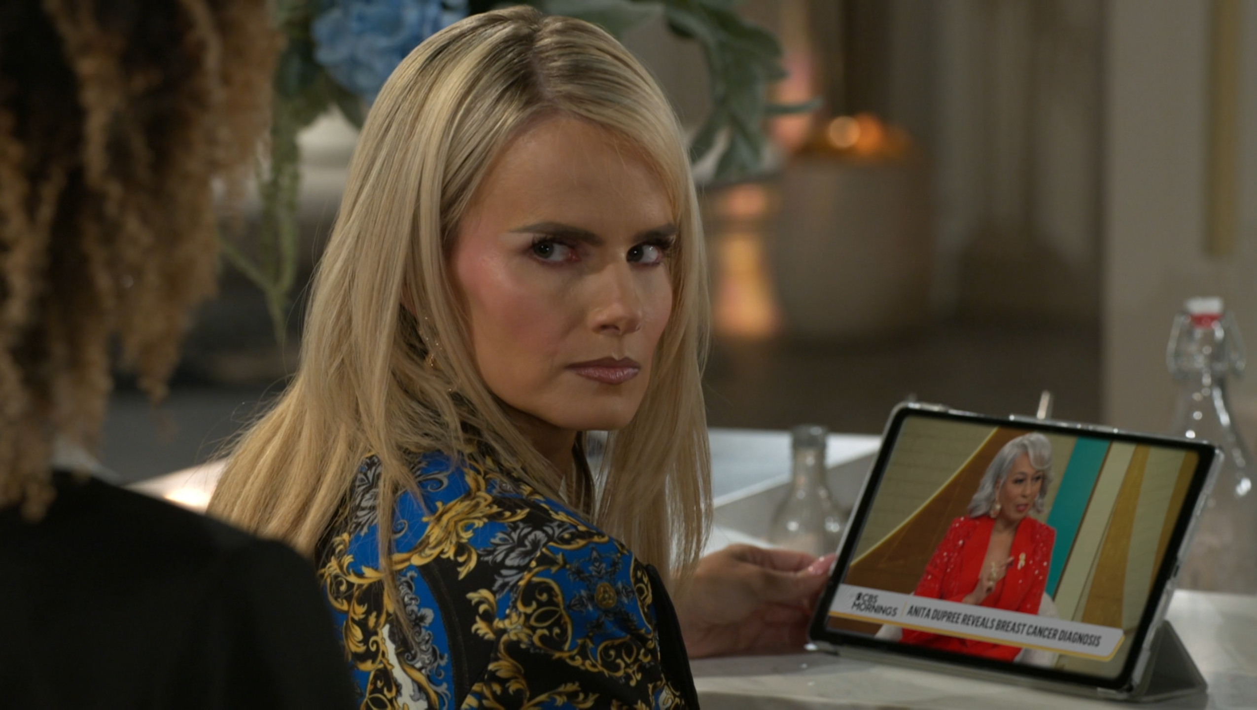 A blonde woman with a serious, intense expression looks over her shoulder toward another person whose back is to the camera, creating a sense of tension or suspicion. She is wearing a dark garment with a bold gold and blue baroque pattern, and her hand rests on a tablet propped up on a white countertop. The tablet displays a news segment from "CBS Mornings" featuring a woman in a red suit with the headline, "Anita Dupree Reveals Breast Cancer Diagnosis." The scene has the polished, high-definition look of a television drama, characterized by shallow depth of field and soft interior lighting that emphasizes the dramatic and somber mood of the interaction.