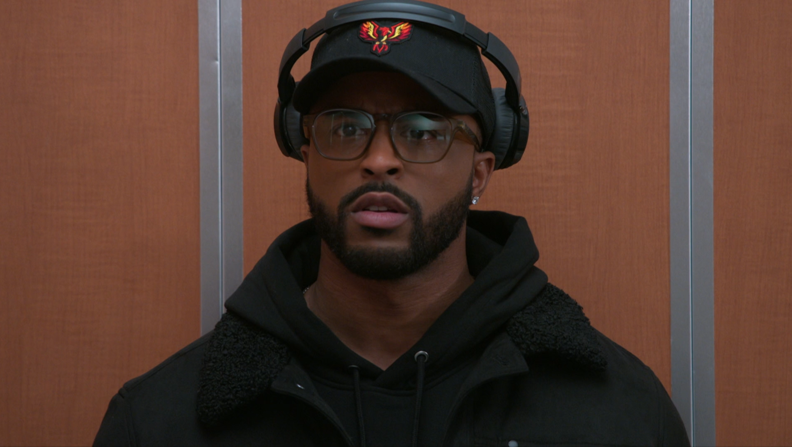 A medium shot of a Black man with a well-groomed beard and mustache, wearing a black baseball cap with a red and orange phoenix logo, black over-ear headphones, and clear-framed glasses. He is dressed in a black hooded sweatshirt under a dark jacket with a textured collar, looking slightly upward with a concerned or bewildered expression, his mouth slightly agape. The background consists of warm, vertical wood paneling separated by thin silver metallic strips, suggesting the interior of an elevator. The lighting is even and soft, giving the image a clean, cinematic feel that captures a moment of quiet tension or unexpected surprise.