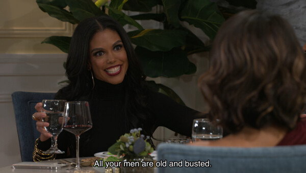 A medium shot from a television scene depicts a woman with long, dark wavy hair and a wide, mischievous grin sitting at a dinner table. She wears a black turtleneck, gold hoop earrings, and numerous gold bangles, holding a water glass in one hand while looking toward another woman whose back is to the camera. The setting is an elegant dining room with a large green plant in the background and a white table set with wine glasses and plates. A subtitle at the bottom reads, 'All your men are old and busted.' The lighting is bright and polished, typical of a modern sitcom or drama, capturing a moment of sharp-tongued, comedic confrontation between friends.