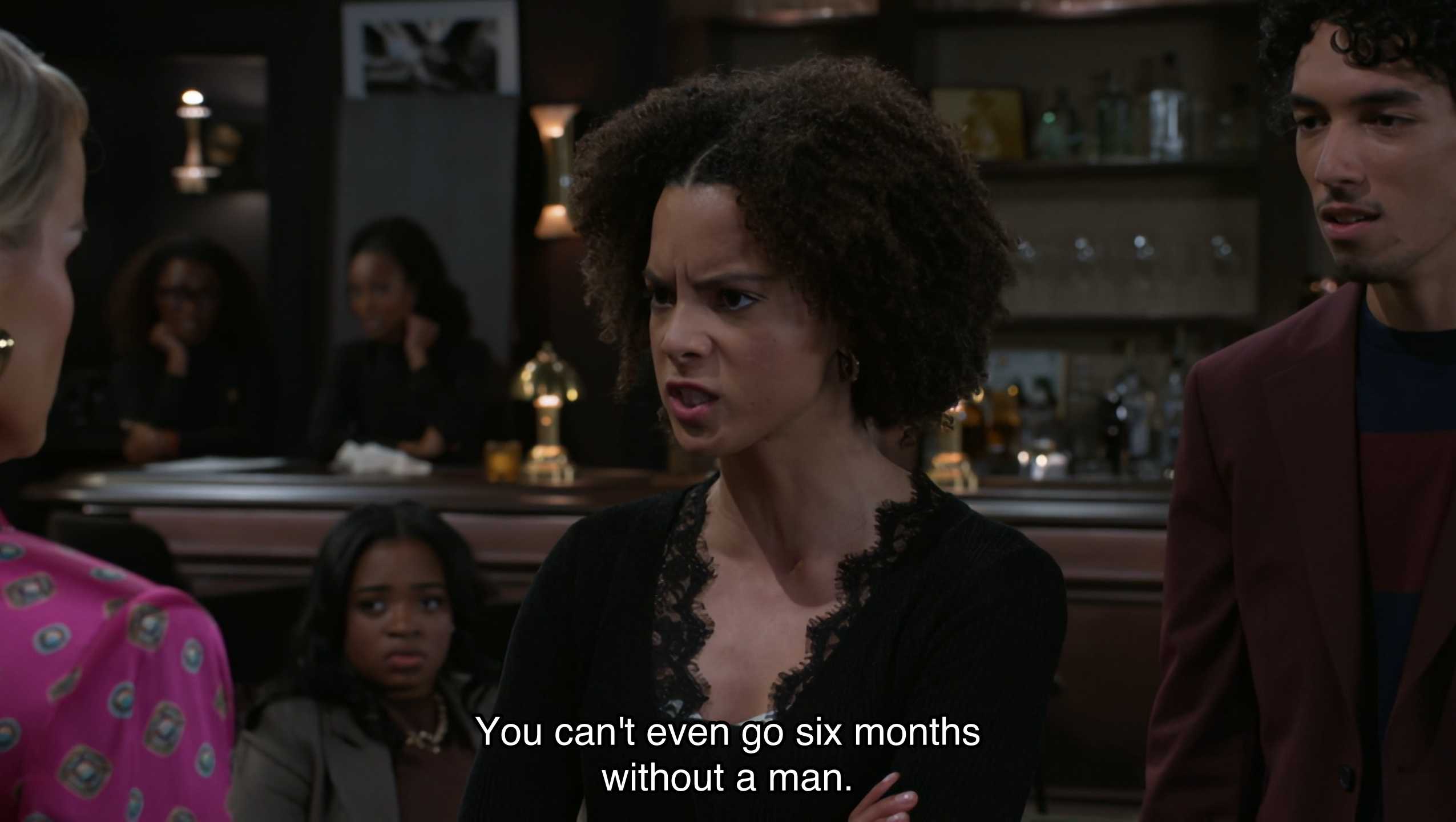 A medium shot from a television series captures an intense confrontation in a dimly lit bar or lounge. Centered is a young woman with curly brown hair, wearing a black lace-trimmed top, her face contorted in an angry, accusatory expression as she speaks to a woman in a pink patterned blouse whose back is partially to the camera. To the right, a young man in a burgundy blazer watches the exchange with a concerned, wide-eyed look, while other patrons in the blurred background observe the drama. A subtitle across the bottom of the frame reads, 'You can't even go six months without a man.' The atmosphere is moody and high-stakes, typical of a dramatic climax in a soap opera or serial drama, emphasized by the low-key lighting and focused character interactions.