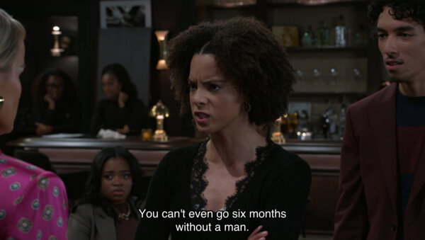 A medium shot from a television series captures an intense confrontation in a dimly lit bar or lounge. Centered is a young woman with curly brown hair, wearing a black lace-trimmed top, her face contorted in an angry, accusatory expression as she speaks to a woman in a pink patterned blouse whose back is partially to the camera. To the right, a young man in a burgundy blazer watches the exchange with a concerned, wide-eyed look, while other patrons in the blurred background observe the drama. A subtitle across the bottom of the frame reads, 'You can't even go six months without a man.' The atmosphere is moody and high-stakes, typical of a dramatic climax in a soap opera or serial drama, emphasized by the low-key lighting and focused character interactions.