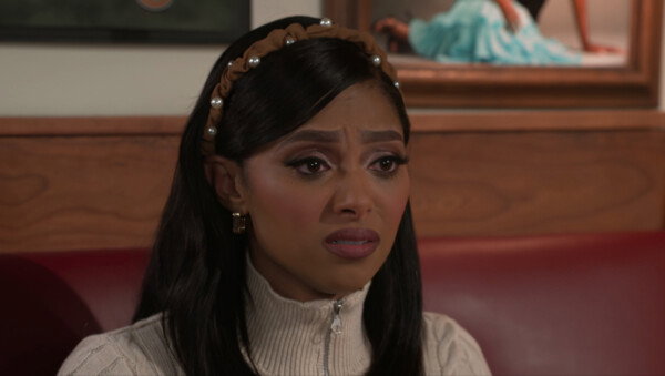 A medium close-up of a young woman with dark hair and a tan, pearl-studded headband shows her with a deeply distressed and pained facial expression. She wears a cream-colored ribbed quarter-zip sweater and small gold hoop earrings, looking slightly off-camera with furrowed brows and her mouth parted in a grimace of concern or sadness. The background features warm wood paneling and a red leather booth, with a framed photograph of a person in a turquoise dress partially visible behind her. The cinematic lighting is soft yet focused, emphasizing her emotional vulnerability and creating a dramatic, high-stakes atmosphere typical of a reality television or soap opera scene.