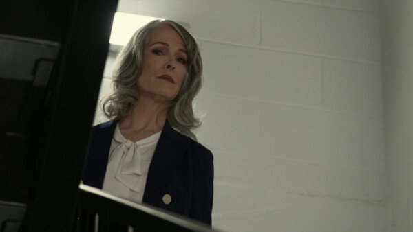 A low-angle, cinematic shot of a mature woman with wavy silver hair looking downward with a stern, authoritative expression. She is dressed in a professional navy blue blazer over a white pussy-bow blouse, positioned against a stark, white cinderblock wall that suggests an institutional or industrial setting. The framing is tight and slightly obscured by a dark foreground element, likely a railing or doorframe, which enhances the sense of observation or confrontation. The lighting is cool and somewhat flat, contributing to a cold, high-stakes, or suspenseful mood that emphasizes the character's formidable presence.