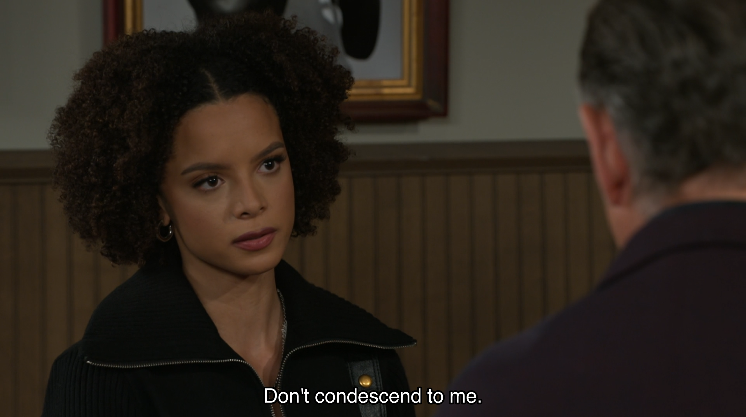 A young woman with curly brown hair and an intense, unwavering gaze looks toward a person whose back is to the camera, her expression one of serious confrontation. She is wearing a black zip-up jacket with a wide collar and small gold hoop earrings, set against a background of wood-paneled walls and a blurred framed picture. White subtitle text at the bottom of the frame reads, 'Don't condescend to me.' The image has the polished, high-definition aesthetic of a modern television drama, characterized by shallow depth of field and soft interior lighting that emphasizes the tense, emotional atmosphere of a personal dispute.