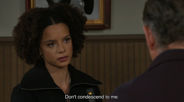 A young woman with curly brown hair and an intense, unwavering gaze looks toward a person whose back is to the camera, her expression one of serious confrontation. She is wearing a black zip-up jacket with a wide collar and small gold hoop earrings, set against a background of wood-paneled walls and a blurred framed picture. White subtitle text at the bottom of the frame reads, 'Don't condescend to me.' The image has the polished, high-definition aesthetic of a modern television drama, characterized by shallow depth of field and soft interior lighting that emphasizes the tense, emotional atmosphere of a personal dispute.