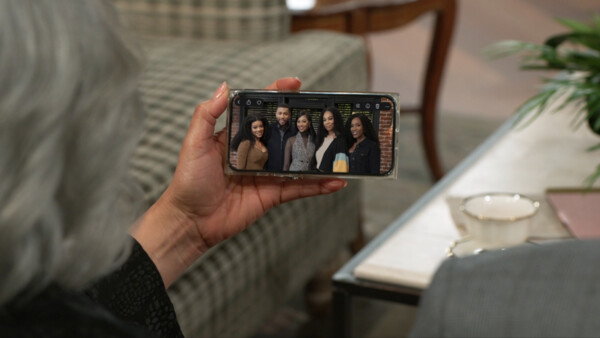 A close-up, over-the-shoulder shot shows a person with gray hair holding a smartphone in their left hand, displaying a horizontal photo of five smiling Black individuals—four women and one man—posed together in an urban setting with a brick wall and window in the background. The person viewing the phone is seated in a cozy, well-lit living room featuring a plaid sofa and a coffee table with a white teacup and green plant, all softly blurred to keep the focus on the digital image. The overall mood is warm, nostalgic, and intimate, captured with a shallow depth of field and soft natural lighting that suggests a reflective moment of personal connection or remembrance.