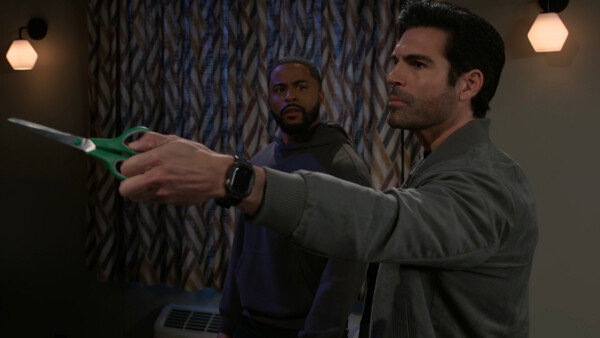 In this medium shot from a television drama, a man with dark hair and a light beard stands in the foreground, pointing a pair of green-handled scissors toward something out of frame with a determined, serious expression. Behind him, another man with a beard and a dark hoodie looks on with a concerned and slightly bewildered gaze. The scene takes place in a dimly lit room featuring neutral-toned walls, glowing hexagonal wall sconces, and a patterned curtain in the background, creating a tense, suspenseful atmosphere. The style is cinematic and realistic, emphasizing a moment of high-stakes confrontation or sudden action between the two characters.