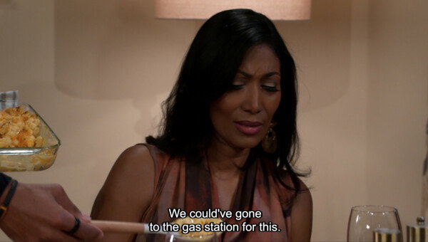 A medium close-up screenshot from a television show or movie features a Black woman with long, dark hair, wearing a sleeveless patterned dress and large earrings, sitting at a dinner table with a distressed or highly disappointed facial expression. Her eyes are lowered and her mouth is downturned as a hand in the foreground holds a serving spoon, and a glass dish of macaroni and cheese is visible on the left side of the frame. White open-captioned subtitles at the bottom center of the screen read, 'We could've gone to the gas station for this.' The overall mood of the scene is comedic yet tense, captured under warm, indoor sitcom-style lighting that emphasizes her humorous expression of disgust during what appears to be a disappointing meal.