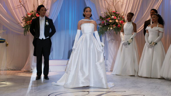 A wide shot from a television show or movie features a formal debutante ball or wedding scene set against an elegant stage with draped white curtains and large floral arrangements. At the center stands a young Black woman looking forward confidently, wearing a voluminous, strapless white satin gown paired with matching over-the-elbow evening gloves. To her left, a young man stands poised in a black tuxedo with tails, a white waistcoat, and a white bow tie. To her right, two other young women stand in similar formal white gowns and long gloves, accompanied by another young man in a dark suit in the background. The overall mood is grand, ceremonial, and highly polished, illuminated by bright, theatrical stage lighting with soft blue undertones that highlights the pristine white attire and the marble-patterned floor.