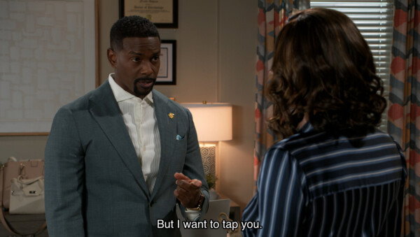 A medium shot from a television drama or sitcom depicts a tense or serious interaction between two characters in a well-lit office setting. On the left, a Black man with a groomed goatee, wearing a tailored grey suit jacket, a white button-down shirt, and a gold lapel pin, is looking intently and pointing a finger toward a woman whose back is to the camera. The woman has shoulder-length, wavy brown hair and is wearing a dark blue and white striped blouse. The background features a softly lit lamp, patterned curtains, and framed certificates on the wall, contributing to a professional yet warm interior aesthetic. White subtitles at the bottom center of the screen read, 'But I want to tap you.'