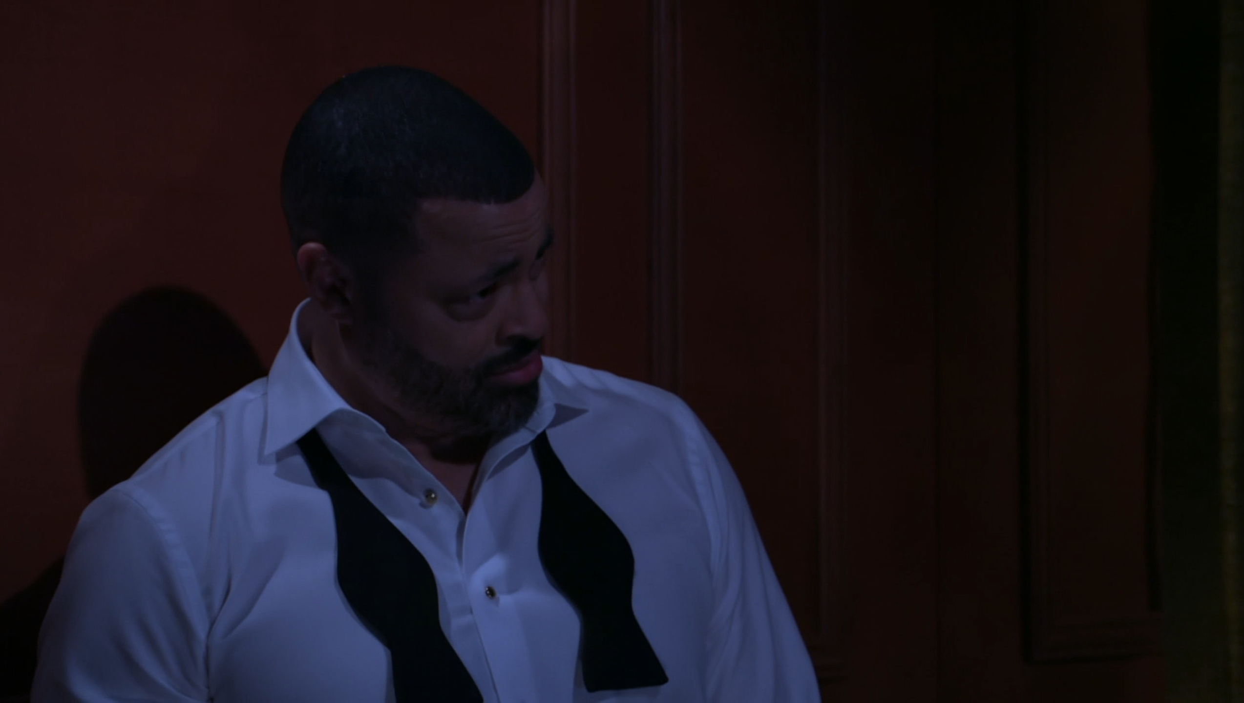 A medium close-up shot from a dramatic production features a man with a short haircut and a trimmed beard, looking downward and to the right with a somber or contemplative expression. He is wearing a white dress shirt with the top buttons undone and an untied, open black bow tie draped around his collar, suggesting the end of a formal event or a moment of distress. The lighting is very dim and moody with a cool blue tint casting over the man, while he stands against a dark, warm-toned wooden paneled wall, enhancing the heavy, emotional atmosphere of the scene.