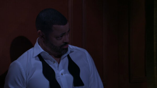 A medium close-up shot from a dramatic production features a man with a short haircut and a trimmed beard, looking downward and to the right with a somber or contemplative expression. He is wearing a white dress shirt with the top buttons undone and an untied, open black bow tie draped around his collar, suggesting the end of a formal event or a moment of distress. The lighting is very dim and moody with a cool blue tint casting over the man, while he stands against a dark, warm-toned wooden paneled wall, enhancing the heavy, emotional atmosphere of the scene.