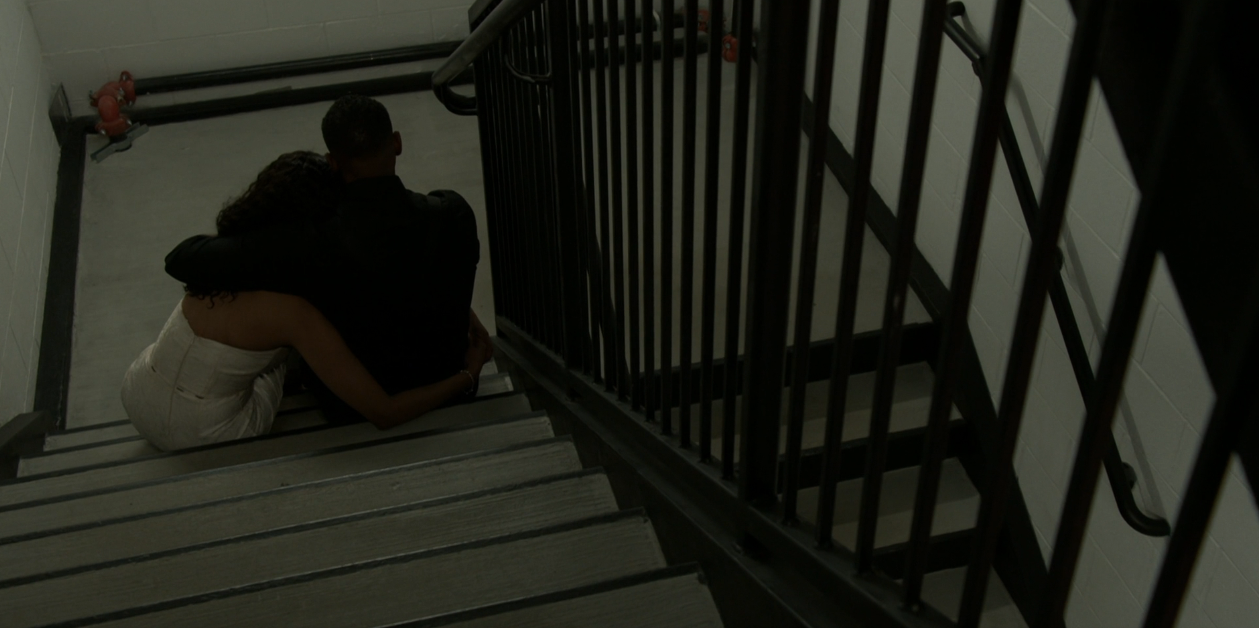 A high-angle shot from a television production shows two people sitting closely together on a concrete landing inside a stark, industrial stairwell, seen from behind. The person on the left is wearing a white dress and leaning into the person on the right, who is dressed entirely in black clothing with an arm wrapped around her shoulders in a comforting gesture. The surrounding environment consists of plain white block walls, dark pipes, and a black metal staircase railing that cuts diagonally across the right side of the frame. The scene is characterized by dim, muted greenish-grey lighting, creating a melancholy, intimate, and secluded atmosphere.