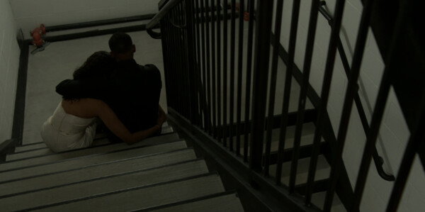 A high-angle shot from a television production shows two people sitting closely together on a concrete landing inside a stark, industrial stairwell, seen from behind. The person on the left is wearing a white dress and leaning into the person on the right, who is dressed entirely in black clothing with an arm wrapped around her shoulders in a comforting gesture. The surrounding environment consists of plain white block walls, dark pipes, and a black metal staircase railing that cuts diagonally across the right side of the frame. The scene is characterized by dim, muted greenish-grey lighting, creating a melancholy, intimate, and secluded atmosphere.
