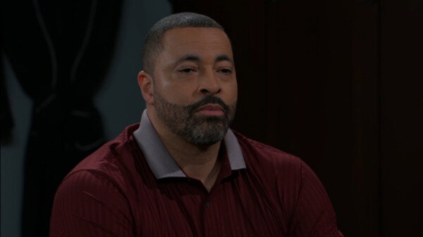 A medium close-up shot from a television drama features a man with a short buzz cut and a neatly trimmed, salt-and-pepper beard. He is wearing a dark maroon polo shirt with a grey collar and appears to be looking straight ahead with a solemn, weary, or contemplative expression. The lighting is low and subdued, with a dark, indistinct background that suggests an indoor evening setting, reinforcing the quiet and serious mood of the scene.