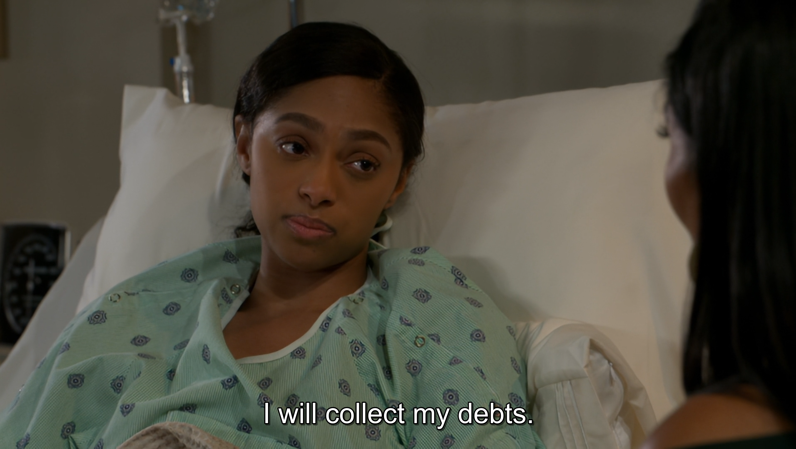 A medium close-up shot from a television drama depicts a young Black woman lying in a hospital bed, propped up against white pillows. She is wearing a light green, patterned hospital gown and has a serious, determined, or skeptical expression as she looks toward another woman whose shoulder and dark hair are visible in the right foreground. The background is slightly out of focus but includes clinical elements like an IV stand and a monitor, contributing to the serious and sterile hospital environment. White subtitles at the bottom center of the frame read, "I will collect my debts."
