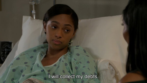 A medium close-up shot from a television drama depicts a young Black woman lying in a hospital bed, propped up against white pillows. She is wearing a light green, patterned hospital gown and has a serious, determined, or skeptical expression as she looks toward another woman whose shoulder and dark hair are visible in the right foreground. The background is slightly out of focus but includes clinical elements like an IV stand and a monitor, contributing to the serious and sterile hospital environment. White subtitles at the bottom center of the frame read, 'I will collect my debts.'