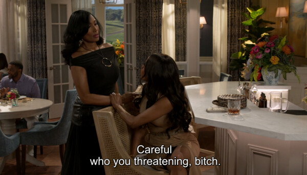 A medium wide shot from a television drama depicts an intense confrontation in an upscale restaurant or country club lounge. On the left, a Black woman with long, straight dark hair stands firmly, wearing an elegant off-the-shoulder black dress and a large gold hoop pendant necklace, while holding onto the arm of another woman seated at the bar. The seated Black woman, seen from a side profile with long wavy dark hair and wearing a beige dress, looks up defiantly at her. The setting is luxurious and brightly lit, featuring an white marble bar top with glassware, a lush, vibrant floral arrangement in the background, open French doors revealing a green landscape outside, and other patrons dining at tables to the far left. White subtitles at the bottom center of the frame read, 'Careful who you threatening, bitch,' reinforcing the tense, high-stakes, and dramatic conflict of the scene.
