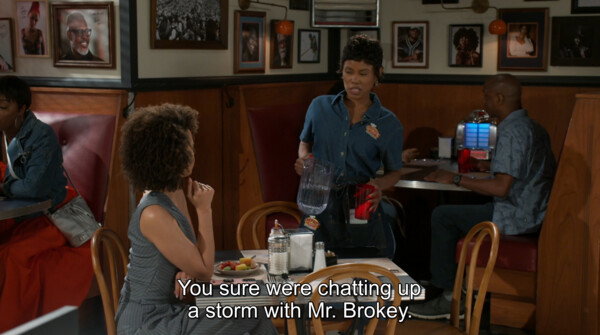 A medium shot from a television show captures a scene inside a brightly lit, classic diner decorated with warm wooden paneling and framed portraits along the wall. In the center, a waitress with short dark hair, dressed in a denim button-down shirt and a black apron, stands at a booth table holding a clear plastic pitcher as she pours water into a red glass. Seated across from her is a Black woman with short, natural afro hair, wearing a sleeveless grey pinstriped top, resting her hand on her chin while listening attentively. On the far right, another customer sits in a booth near a vintage jukebox. The lighting is warm and natural for a daytime indoor setting, enhancing the casual and conversational mood. White subtitles at the bottom center of the screen read, 'You sure were chatting up a storm with Mr. Brokey.'