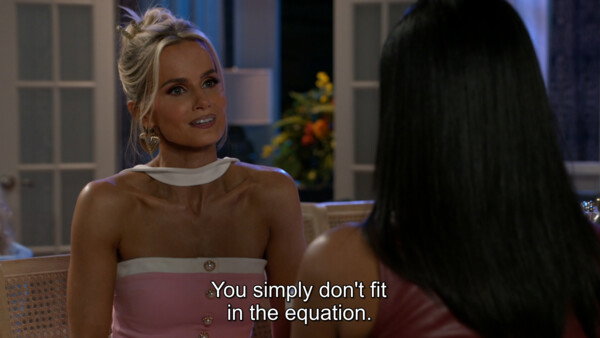 A over-the-shoulder medium shot from a television drama features a blonde woman with her hair styled in an updo and heart-shaped gold earrings, looking intently at another woman whose long dark hair fills the right foreground. The blonde woman is wearing a strapless pink and white dress with pearl buttons, paired with a white neck band, and carries a subtle, smug, or confrontational expression. Set in an elegant, soft-lit indoor room with french doors and floral accents in the background, the scene conveys a tense and dramatic atmosphere. White subtitles at the bottom center read, 'You simply don't fit in the equation.'