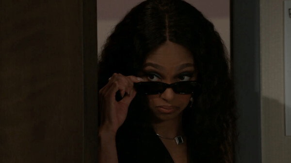 A close-up shot from a television production shows a Black woman with long, dark, wavy hair peering around the edge of a doorway or partition. She lowers a pair of slim black sunglasses with one hand to peer intently over the top rim, sporting a skeptical, suspicious, or secretive expression. She wears dark eyeshadow, a dark outfit, and a subtle necklace with a square pendant. The scene is lit with low, moody lighting that casts shadows around the entryway, creating a sense of drama, suspense, and stealthy observation.
