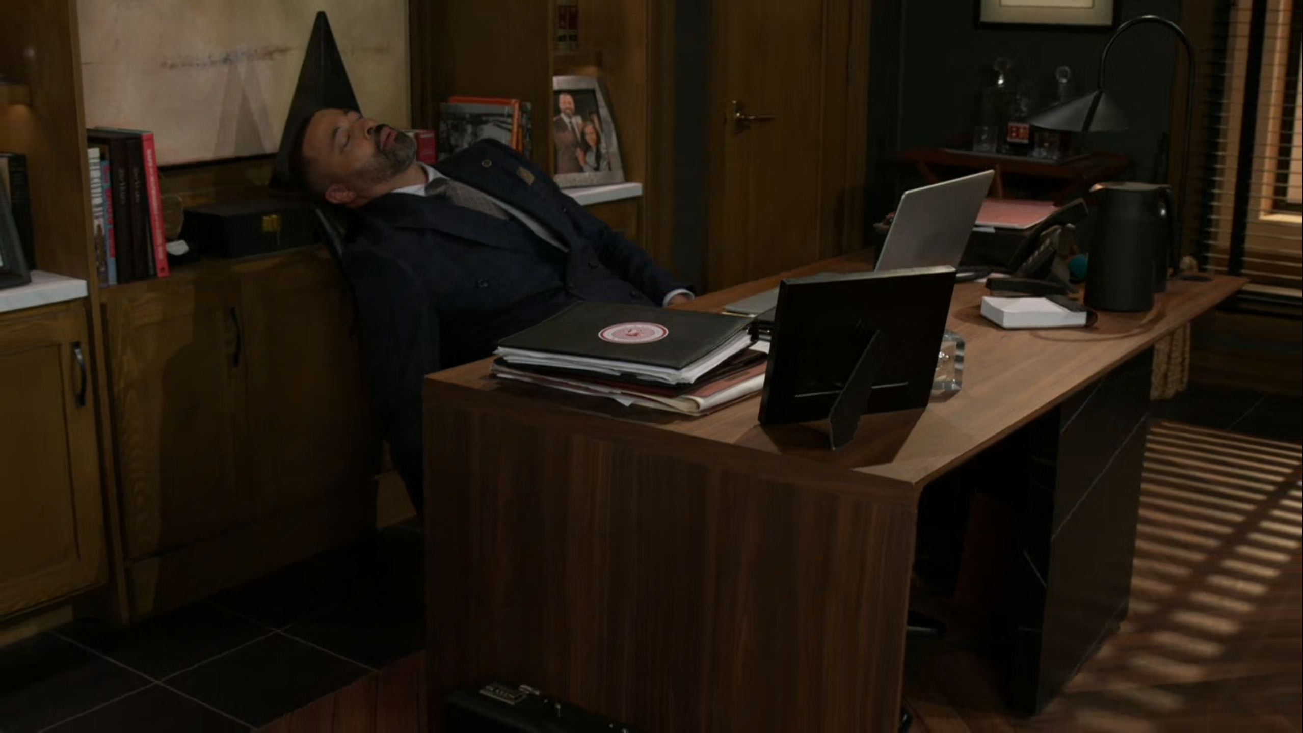A medium wide shot from a television production depicts a man in a dark double-breasted suit leaning far back in his executive desk chair with his head tilted back, either asleep or unconscious. He sits behind a large wooden office desk cluttered with stacked folders, a black picture frame, an open laptop, and a dark pitcher. The warm, dimly lit office features dark wood cabinetry, bookshelves filled with framed photos and books, and shadows cast across the floor by window blinds, creating a quiet, moody, and dramatic tone suggestive of exhaustion, tension, or distress.
