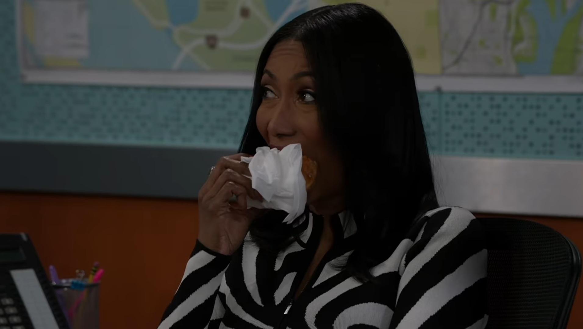 A close-up shot from a television drama features a Black woman with long, sleek dark hair, taking a bite of a pastries or sandwich wrapped in a white paper napkin. She glances upward and off-camera to the left with wide, playful, or secretive eyes, conveying a sense of sly amusement. She wears a bold, black-and-white graphic zebra-striped cardigan sweater. The background shows an office setting with maps framed on the wall behind her, presented under soft, slightly dimmed interior studio lighting that creates a candid, lighthearted, and engaging dramatic moment.