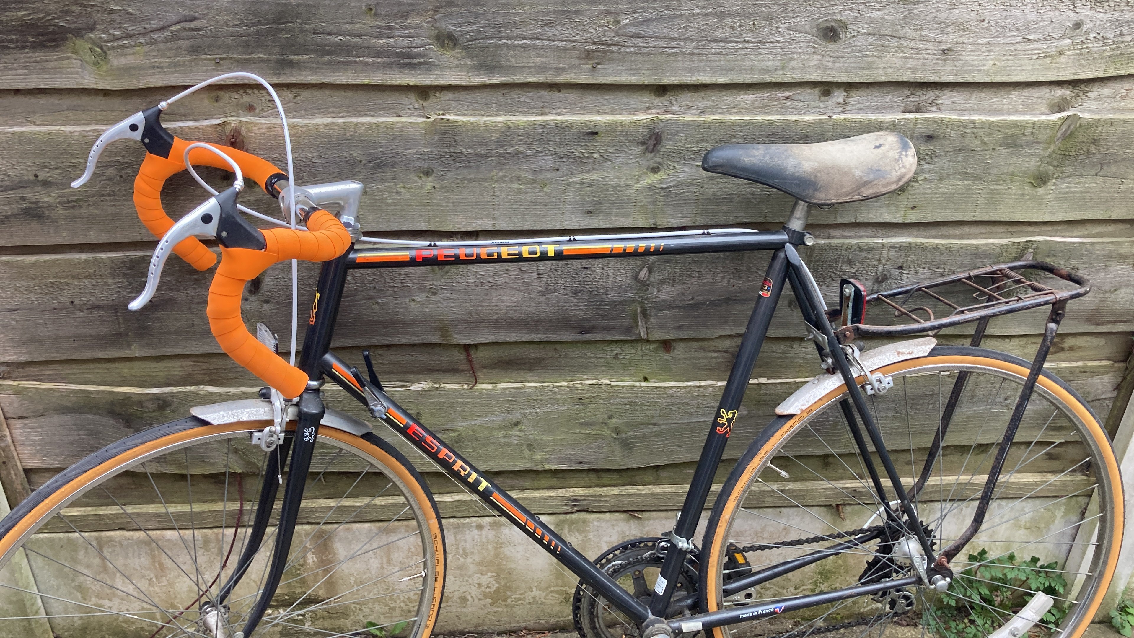 A vintage Peugeot bike  leaning against a fence. It has orange handlebars, and a black frame, with extremely-80's red-yellow gradient text on the frame.