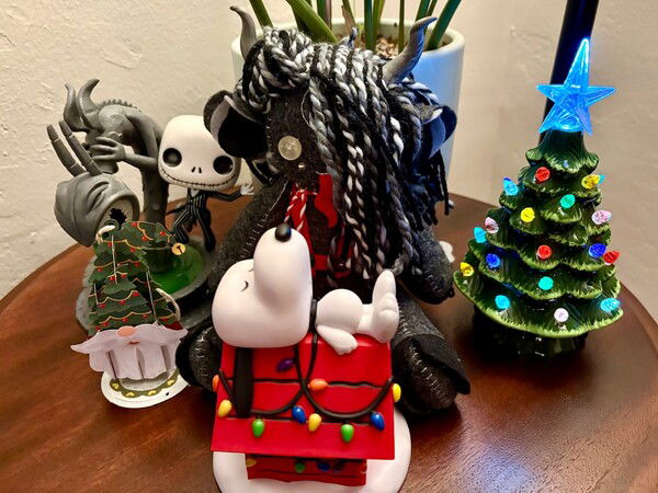 A collection of figurines on a wooden table, featuring a plush Krampus, a Funko Pop Jack Skellington, a paper pop-up Santa Claus wearing a Christmas tree hat, a Funko Pop Snoopy sleeping on his bright red doghouse adorned with Christmas lights, and a small lit ceramic Christmas tree.