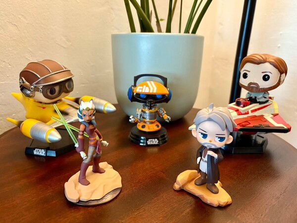 Star Wars figures arranged on a dark wood table in front of a potted plant, from left to right: Funko Pop of Anakin Skywalker with R2-D2 in a Naboo Starfighter, Ahsoka Tano, Funko Pop of DJ Rex, Obi-Wan Kenobi, and a Funko Pop of Obi-Wan Kenobi in a Delta 7 Jedi Starfighter.