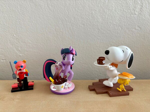Three figures, from left to right, a bear costume Lego minifig with a peg leg and coffee mug, a Twilight Sparkle figure with a coffee mug, and a Snoopy figure with a coffee mug on a plate followed by Woodstock with a plate of pancakes.