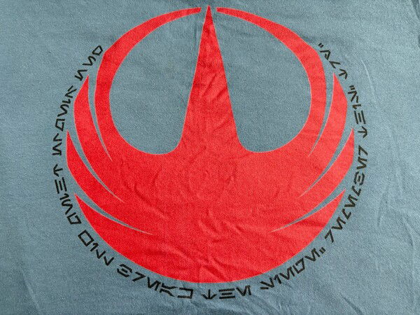 Photo of a blue shirt with the Star Wars Rebel Alliance symbol and Aurebesh text that translates to: “One single thing will break the siege. Remember this: Try.”