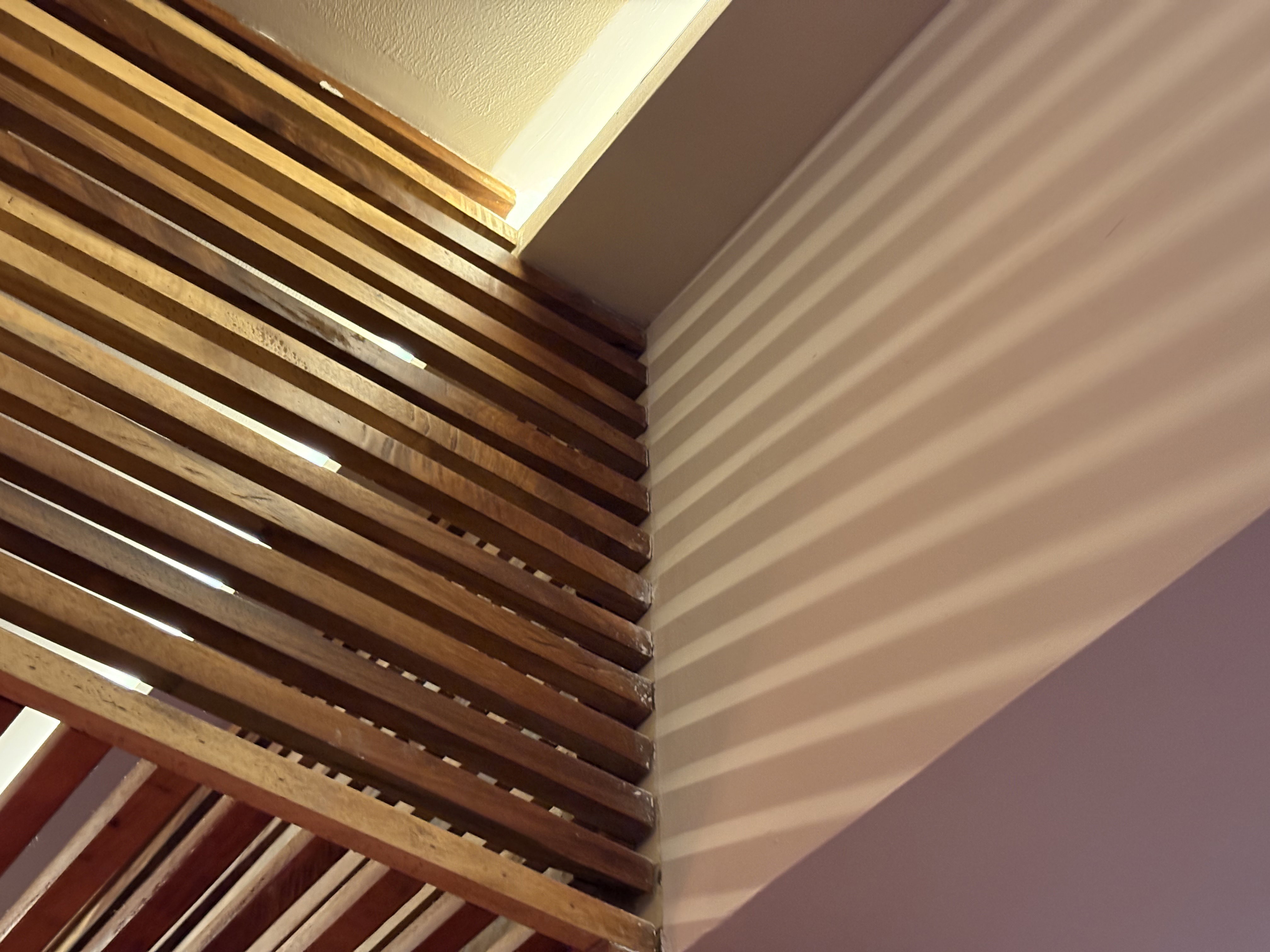 Close-up of wooden horizontal slats forming a room divider or decorative wall feature, meeting a white ceiling with crown molding. The warm-toned wooden slats create interesting shadow patterns on the adjacent white wall. The wooden element appears to be a custom architectural detail that combines modern slatted design with traditional interior elements, creating a play of light and texture in a restaurant space.​​​​​​​​​​​​​​​​