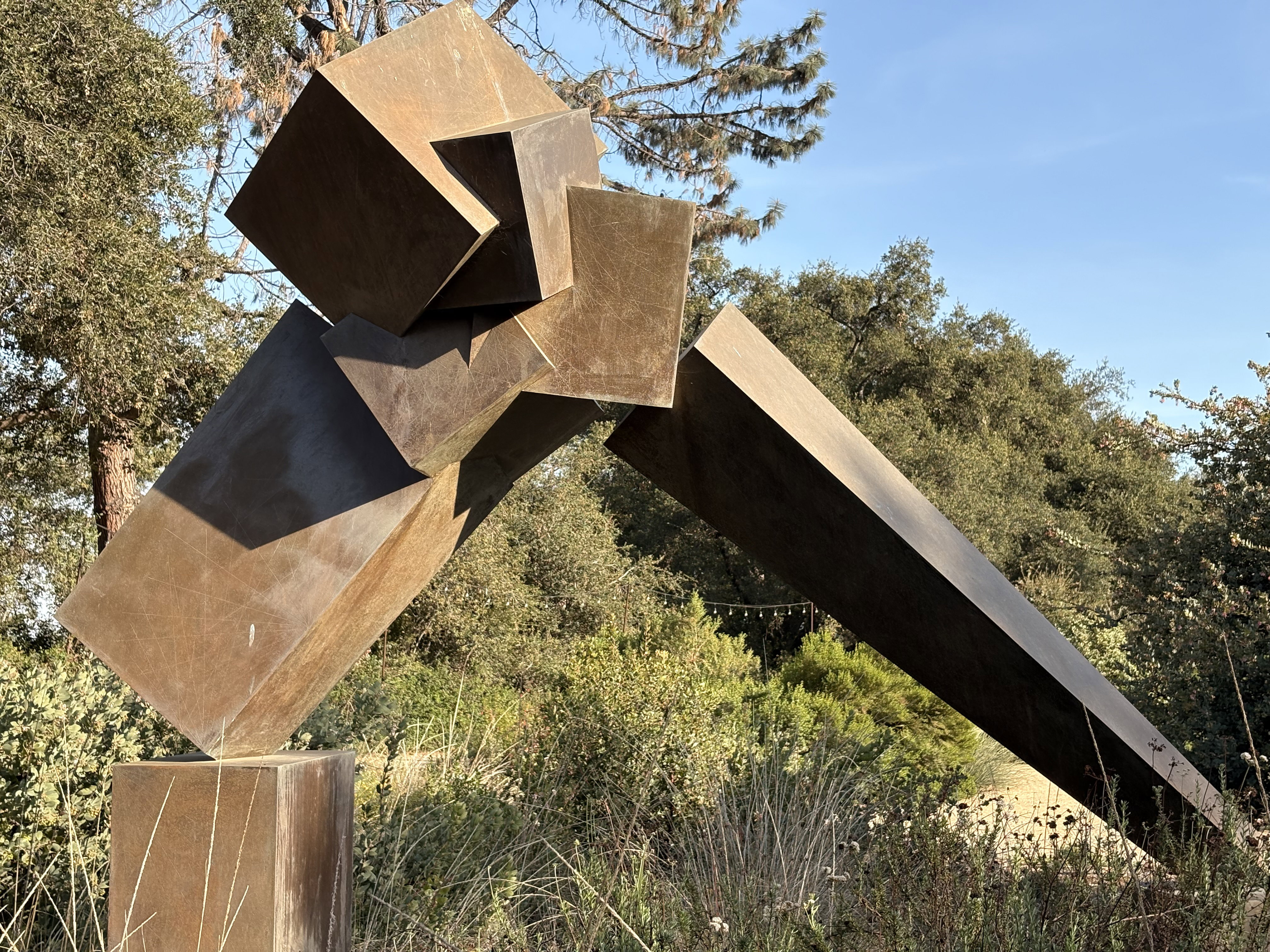 This outdoor sculpture features several geometric copper or bronze-colored metal cubes and rectangular forms arranged in a dynamic, angular composition. The sculpture projects outward with multiple rectangular pieces intersecting at various angles. Set against a backdrop of trees and vegetation with a blue sky visible, the metallic surfaces show weathering and patina. The abstract arrangement creates a sense of movement and tension through the balanced yet seemingly precarious positioning of the geometric forms.​​​​​​​​​​​​​​​​