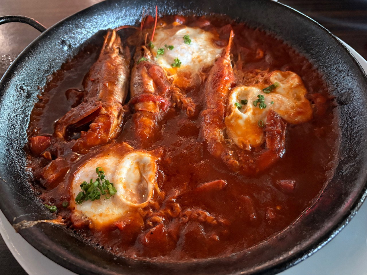 A cast-iron skillet containing a rich, red tomato-based stew with seafood. Several prawns with their heads intact are visible in the sauce. Poached eggs are scattered throughout, garnished with small green herbs. The dish has a rustic, homemade appearance.