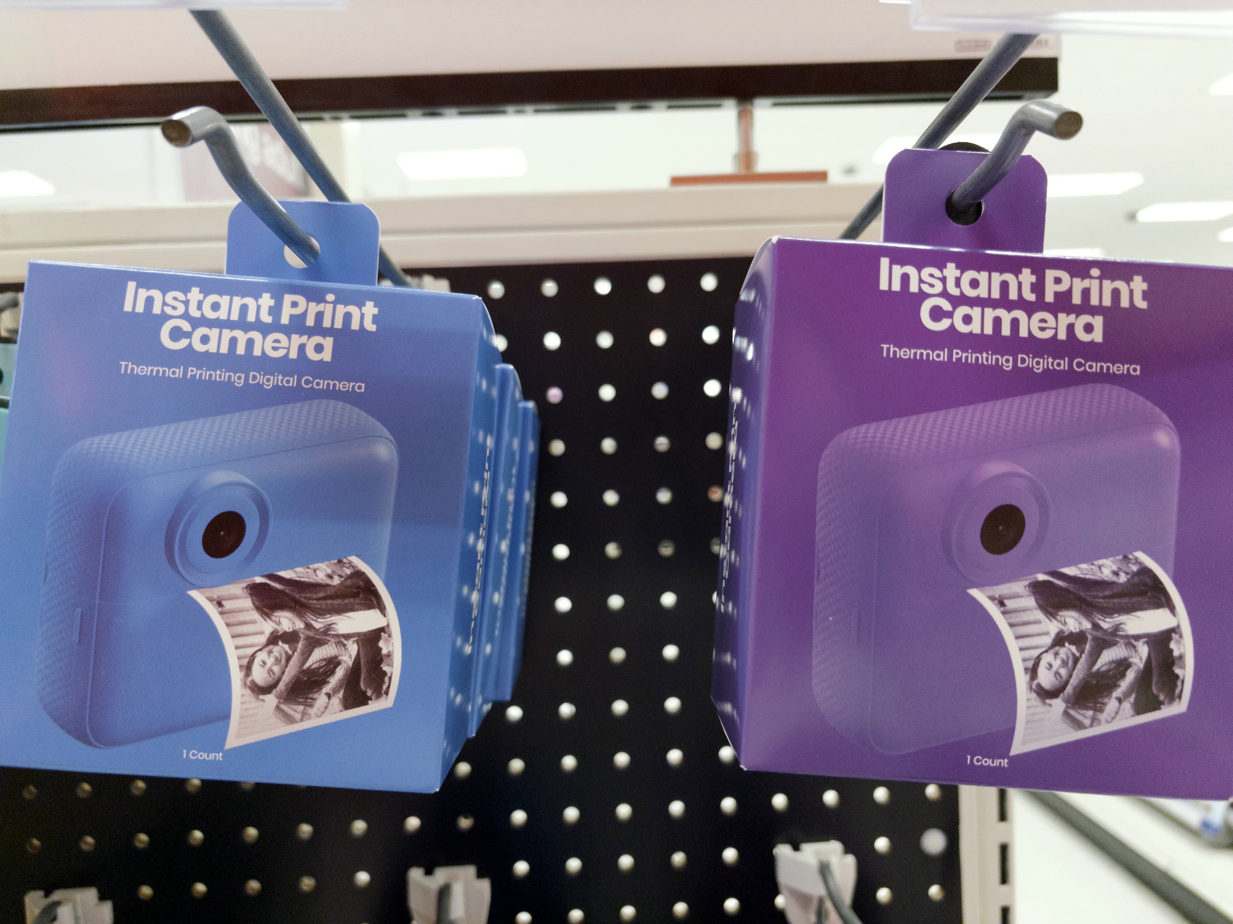 Two hanging boxes of cameras, one blue and one purple, labeled “Instant Print Camera - Thermal Printing Digital Camera.”