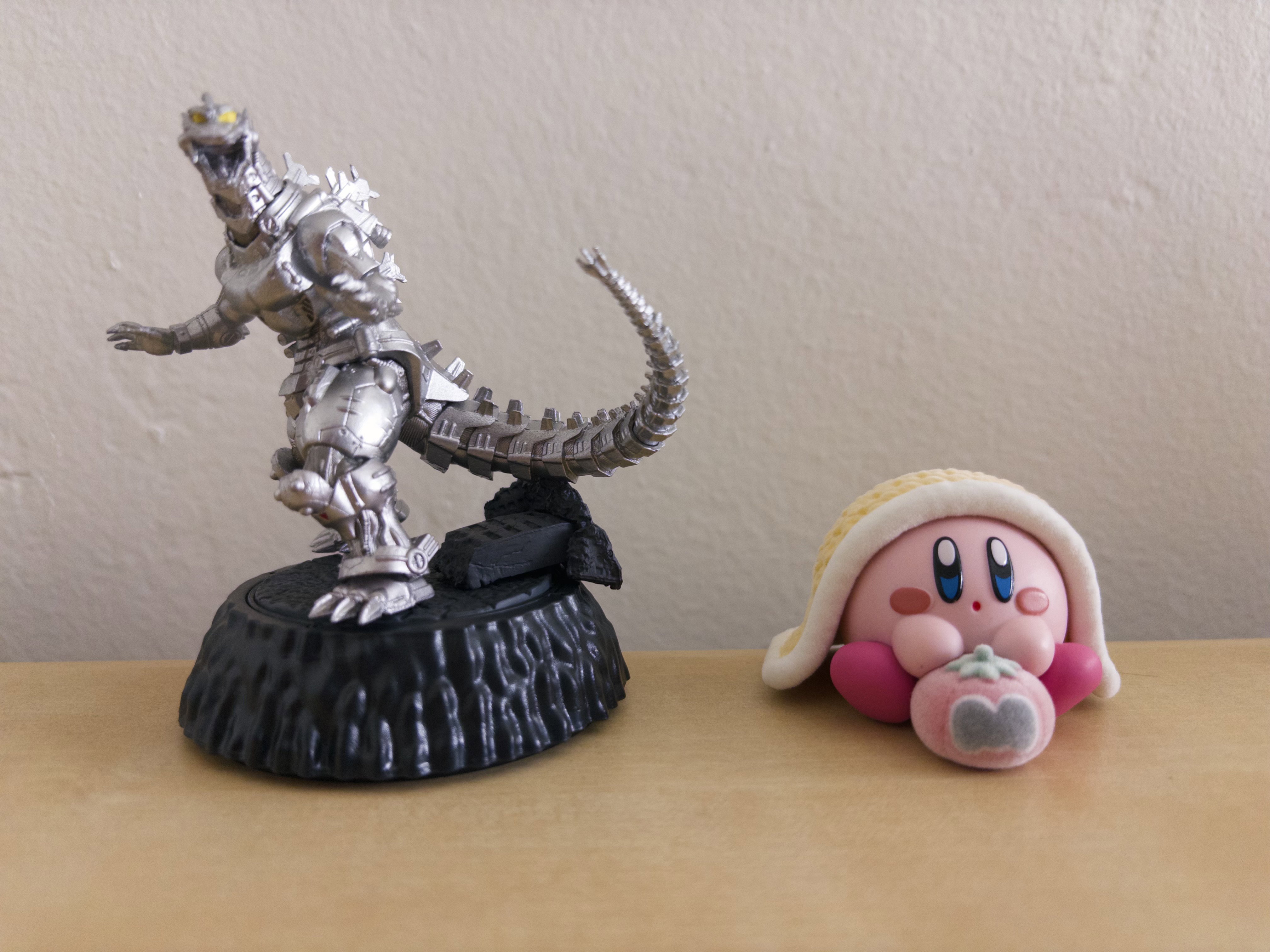 A small Mechagodzilla figure in a defiant post is positioned next to a small Kirby figure under a yellow blanket.